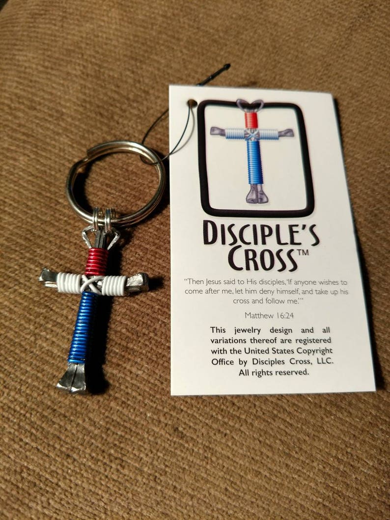 Puede incluir: Un llavero con forma de cruz de clavos de herradura rojo, blanco y azul con un anillo plateado. El llavero est&aacute; unido a una tarjeta con el texto "Disciple's Cross" y una cita de Mateo 16:24. La tarjeta tambi&eacute;n indica que el dise&ntilde;o est&aacute; registrado en la Oficina de Derechos de Autor de los Estados Unidos por Disciples Cross, LLC.