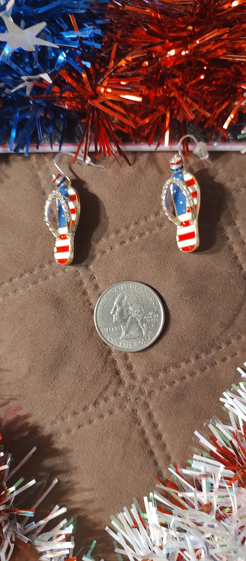 Puede incluir: Un par de pendientes colgantes de tono dorado con un dise&ntilde;o de chanclas rojo, blanco y azul con pedrer&iacute;a. Los pendientes est&aacute;n sobre una superficie marr&oacute;n con oropel rojo, blanco y azul.