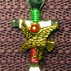 May include: A gold eagle with outstretched wings is attached to a cross made of wire wrapped in green, red, and white thread. The cross is attached to a black cord.