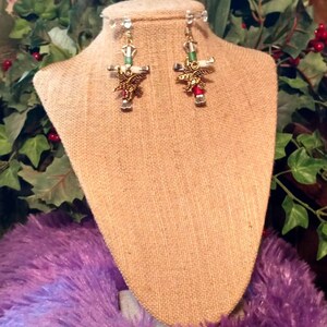 May include: A pair of gold-tone earrings with a cross design and a small bird charm. The earrings are hanging from a display stand.