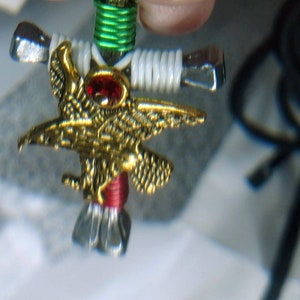 以下が含まれることがあります： 赤い宝石が付いた金色のワシのデザインの手作りのペンダント。ペンダントは黒いコードに付けられています。ワシは金属製の十字架に取り付けられ、白と緑のワイヤーで包まれています。