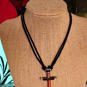 May include: A handmade necklace featuring a cross pendant. The cross is crafted from silver-toned metal and wrapped with red wire. The necklace has a black cord.