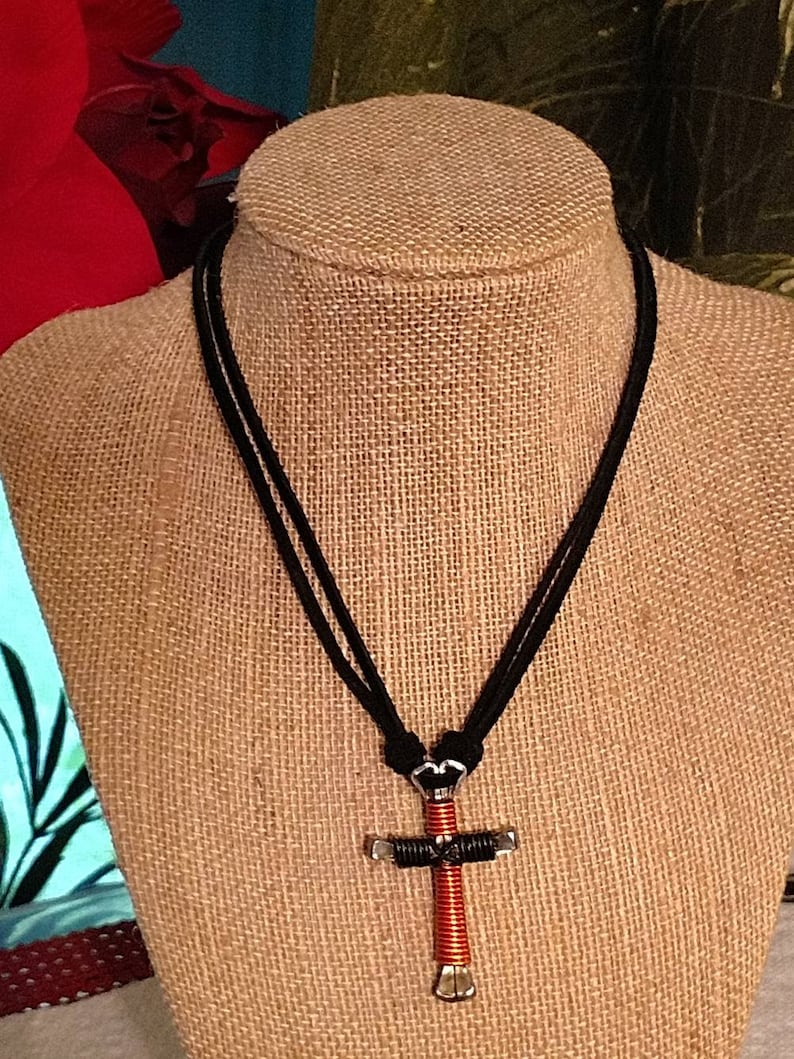 May include: A handmade necklace featuring a cross pendant. The cross is crafted from silver-toned metal and wrapped with red wire. The necklace has a black cord.