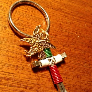 以下が含まれることがあります： 金色のワシのペンダントが付いた手作りのキーホルダー。キーホルダーの十字型のデザインは、赤、白、緑の巻き線と金属製の金具を組み合わせています。キーリングは銀色です。