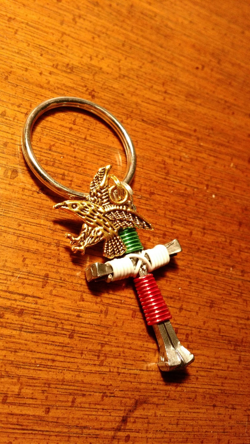 以下が含まれることがあります： 金色のワシのペンダントが付いた手作りのキーホルダー。キーホルダーの十字型のデザインは、赤、白、緑の巻き線と金属製の金具を組み合わせています。キーリングは銀色です。