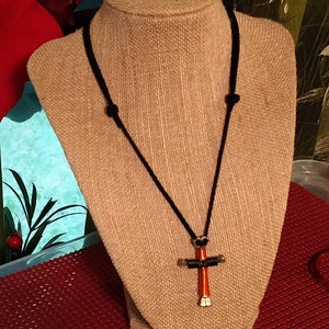 May include: A handmade necklace featuring a cross pendant. The cross is crafted from silver-colored metal and wrapped with orange wire. It hangs from a black cord necklace, displayed on a tan mannequin.