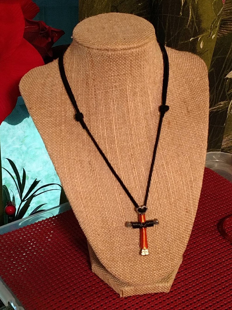 May include: A handmade necklace featuring a cross pendant. The cross is crafted from silver-colored metal and wrapped with orange wire. It hangs from a black cord necklace, displayed on a tan mannequin.