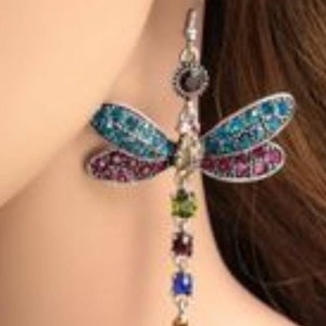 May include: A pair of dangle earrings featuring a dragonfly design with blue, green, and purple rhinestones. The dragonfly is attached to a chain with colorful beads.