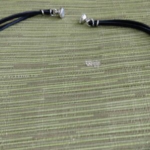 Op de afbeelding: Een zwarte leren choker ketting met zilveren metalen sluitingen. De ketting ligt plat op een groen, getextureerd oppervlak.