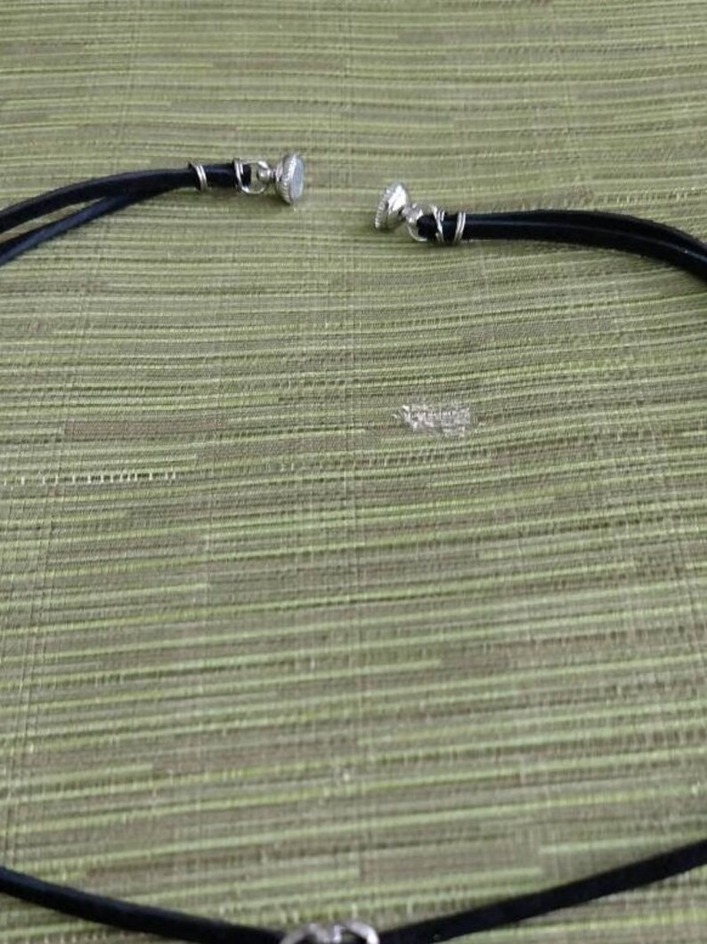 Op de afbeelding: Een zwarte leren choker ketting met zilveren metalen sluitingen. De ketting ligt plat op een groen, getextureerd oppervlak.