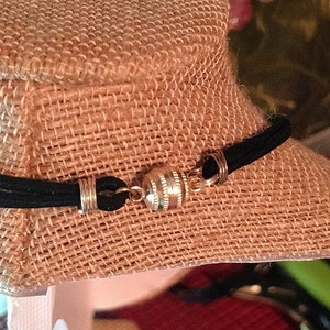 May include: A black necklace with a magnetic clasp, displayed on a burlap-covered bust. The clasp is silver and cylindrical, connecting the ends of the necklace. The necklace appears to be made of a black, suede-like material.