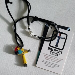 May include: A yellow and black wire cross with a blue, green, and red puzzle piece heart pendant. The cross is on a black cord necklace. The tag reads "Disciple's Cross" and includes a quote from Matthew 16:24. "Then Jesus said to His disciples, "If anyone wishes to come after me, let him deny himself, and take up his cross and follow me."  Matthew 16:24