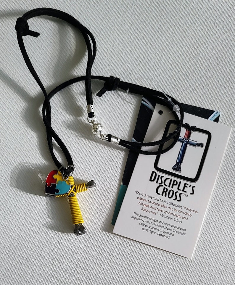 May include: A yellow and black wire cross with a blue, green, and red puzzle piece heart pendant. The cross is on a black cord necklace. The tag reads "Disciple's Cross" and includes a quote from Matthew 16:24. "Then Jesus said to His disciples, "If anyone wishes to come after me, let him deny himself, and take up his cross and follow me."  Matthew 16:24