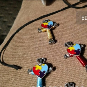 May include: Three colourful puzzle piece heart charms on a necklace. Each charm is made of metal and has a different coloured puzzle piece on a heart-shaped background. The charms are attached to a black cord.