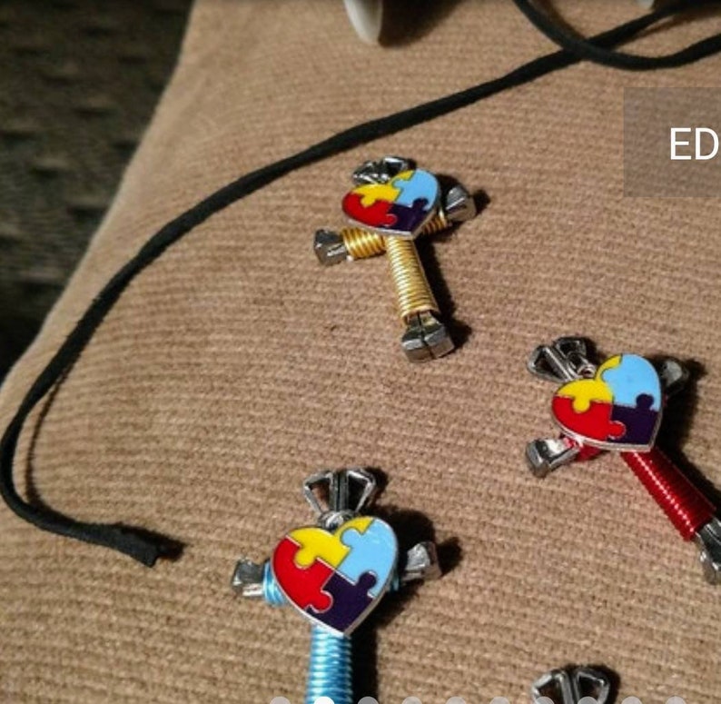 May include: Three colourful puzzle piece heart charms on a necklace. Each charm is made of metal and has a different coloured puzzle piece on a heart-shaped background. The charms are attached to a black cord.