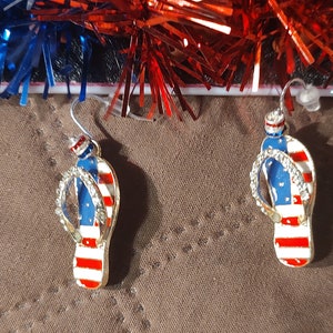 Puede incluir: Un par de pendientes de color dorado en forma de chanclas con esmalte rojo, blanco y azul, que se asemejan a la bandera estadounidense.