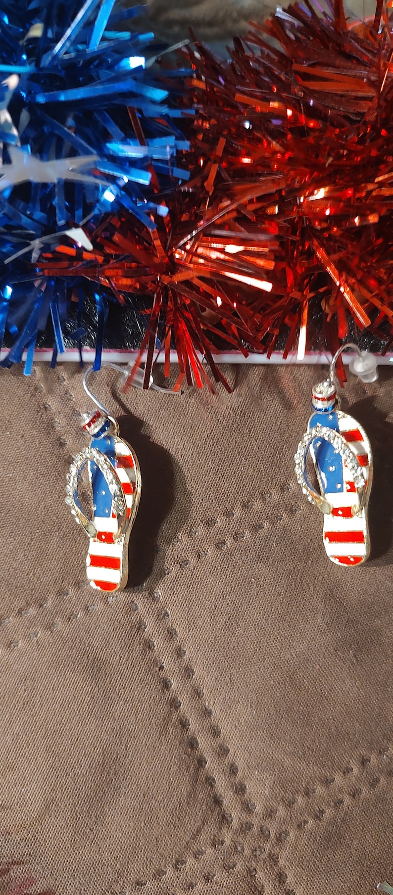 Puede incluir: Un par de pendientes de color dorado en forma de chanclas con esmalte rojo, blanco y azul, que se asemejan a la bandera estadounidense.