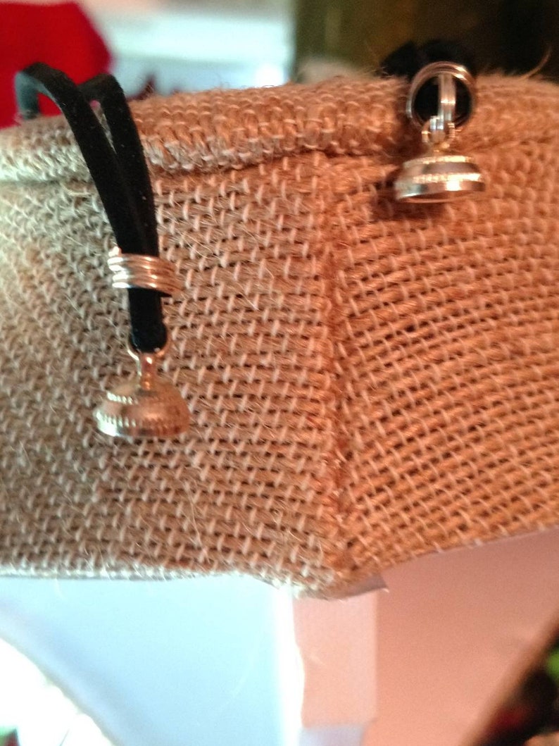 May include: Close-up of two small, silver-colored pendants with a circular design, attached to black cords. The pendants are hanging against a textured, tan fabric background. The pendants have a decorative, detailed design.