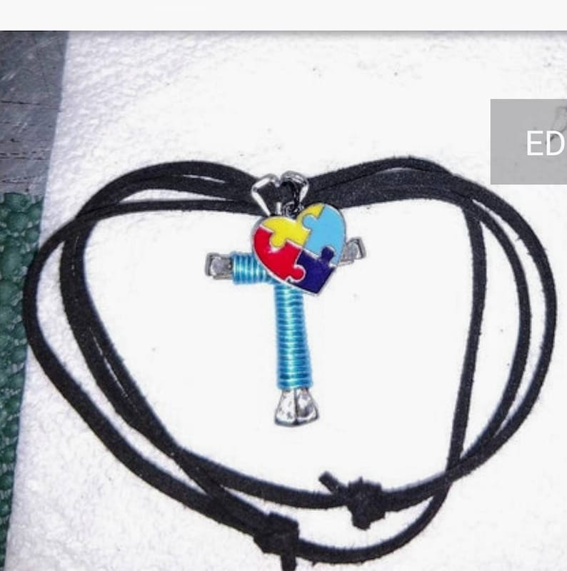 May include: A black cord necklace with a silver cross pendant. The cross is decorated with a blue, red, yellow and purple puzzle piece heart.