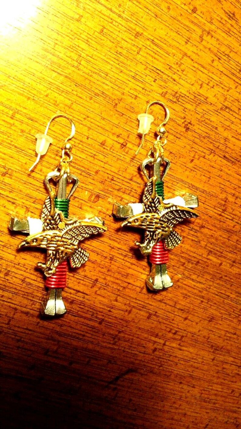 May include: A pair of silver and gold earrings featuring a cross with a red, green, and white wrapped nail, topped with a golden eagle.