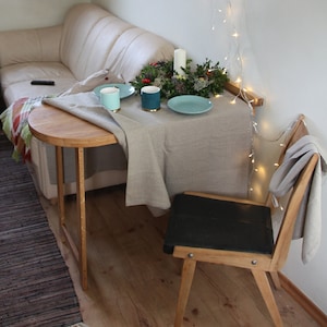 May include: A small, round wooden table with a white tablecloth set for two. There are two blue mugs, a green wreath with candles, and a blue plate on the table. A wooden chair with a black seat sits next to the table. The table is in a living room with a couch and a window in the background.