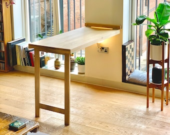 Könnte beinhalten: Ein heller Holz-Klapptisch mit schlichtem Design, an einer weißen Wand befestigt. Der Tisch steht neben einem Fenster mit Blick auf ein Backsteingebäude.