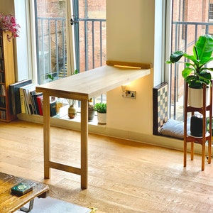 May include: A light wood folding table with a simple design, attached to a wall with a white surface. The table is positioned next to a window with a view of a brick building.