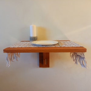 May include: A wooden wall-mounted table with a white plate, a fork, and a white candle on a lace tablecloth.