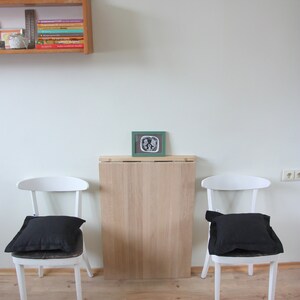May include: A minimalist interior scene featuring two white chairs with black cushions flanking a light wood table. A framed picture and a shelf with books and a photo are mounted on the white wall. The floor is light wood.