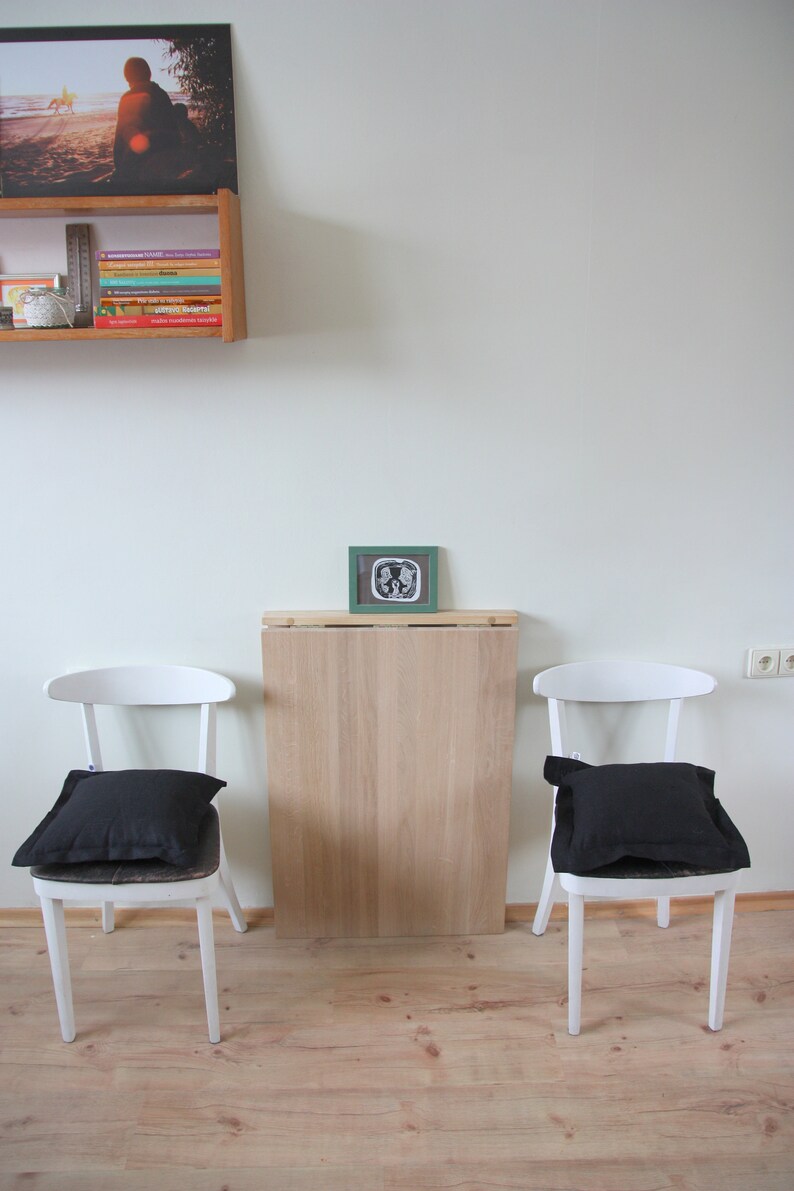 May include: A minimalist interior scene featuring two white chairs with black cushions flanking a light wood table. A framed picture and a shelf with books and a photo are mounted on the white wall. The floor is light wood.