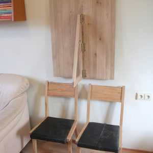 Puede incluir: Dos sillas de madera con asientos de tela negra se encuentran frente a una mesa de madera montada en la pared que se pliega hacia abajo. La mesa est&aacute; unida a la pared con bisagras.