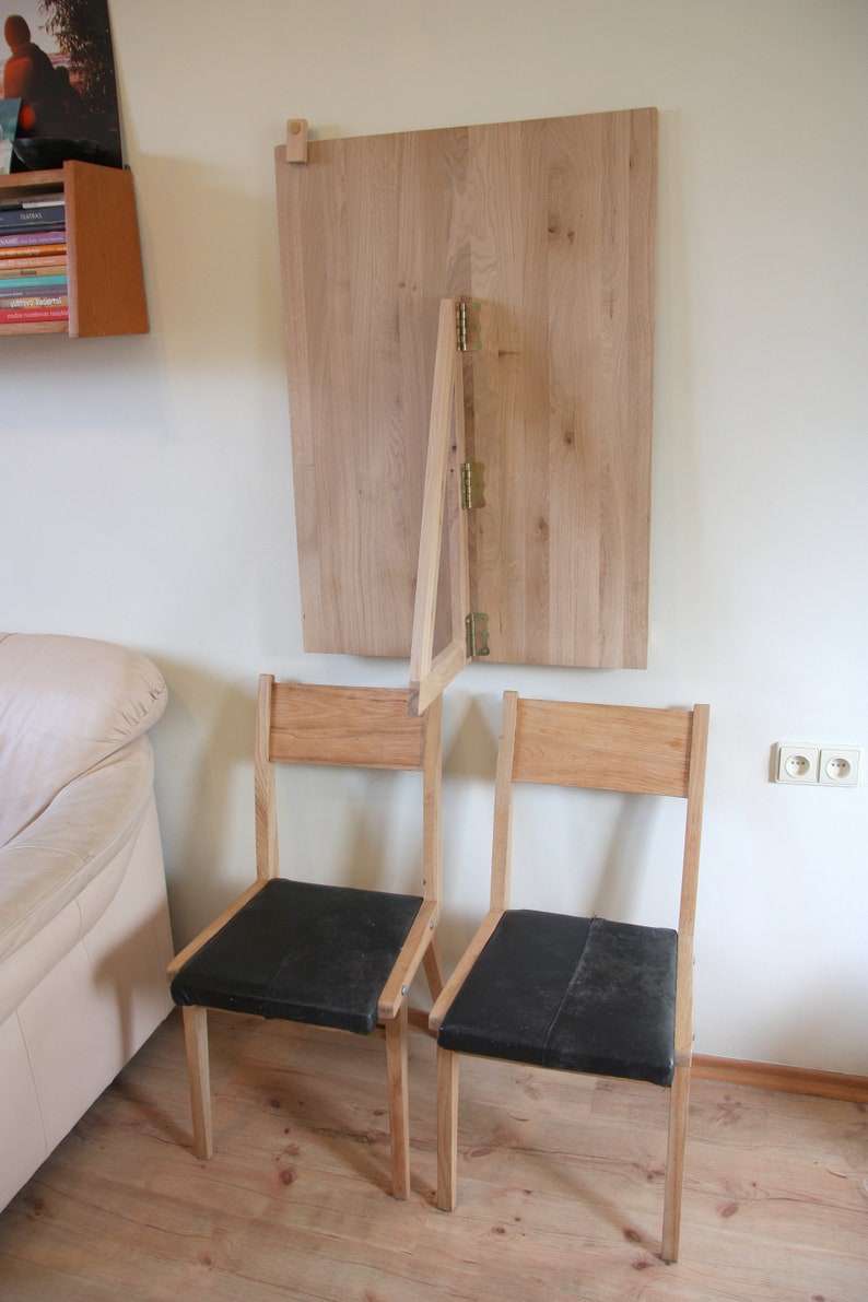 Puede incluir: Dos sillas de madera con asientos de tela negra se encuentran frente a una mesa de madera montada en la pared que se pliega hacia abajo. La mesa est&aacute; unida a la pared con bisagras.