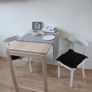 May include: A small, light-colored wooden table with a gray runner, set with a white plate and a mug. Two white chairs, one with a black cushion, are positioned on either side. A small green framed picture and books are on the table.