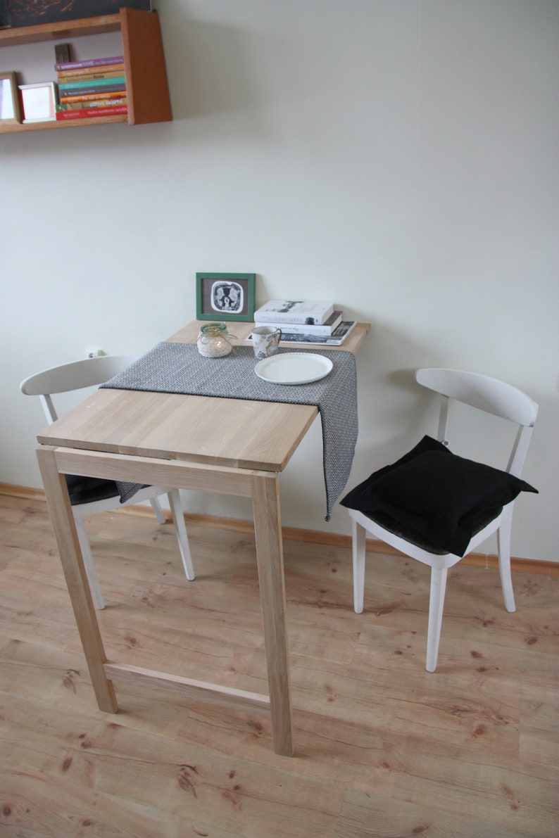 May include: A small, light-colored wooden table with a gray runner, set with a white plate and a mug. Two white chairs, one with a black cushion, are positioned on either side. A small green framed picture and books are on the table.