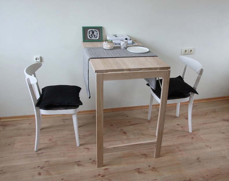 May include: A small, light-colored wooden table with a gray runner, a white plate, and a small decorative item. Two white chairs with black cushions are positioned on either side of the table. A framed picture rests on the table.