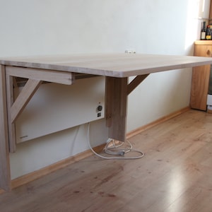 May include: A light wood folding table mounted to a wall with a white electric heater underneath. The table is made of unfinished wood and has a simple design.