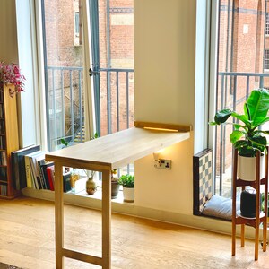 May include: A light wood folding desk with a white wall behind it. The desk is attached to the wall and has a light strip underneath it. The desk is in a room with a window and a plant.