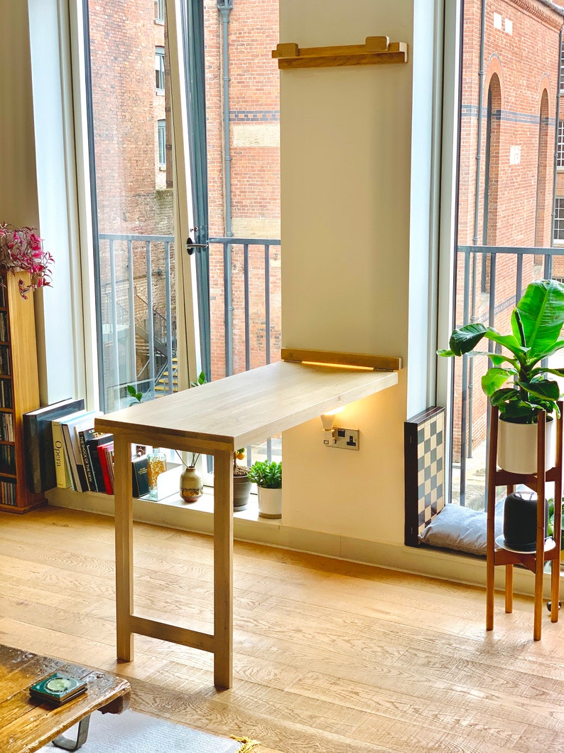 May include: A light wood folding desk with a white wall behind it. The desk is attached to the wall and has a light strip underneath it. The desk is in a room with a window and a plant.