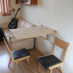 Puede incluir: Una mesa plegable de madera clara con sillas negras est&aacute; montada en una pared. La mesa est&aacute; plegada y no est&aacute; en uso. Las sillas son de madera clara con asientos de cuero negro.