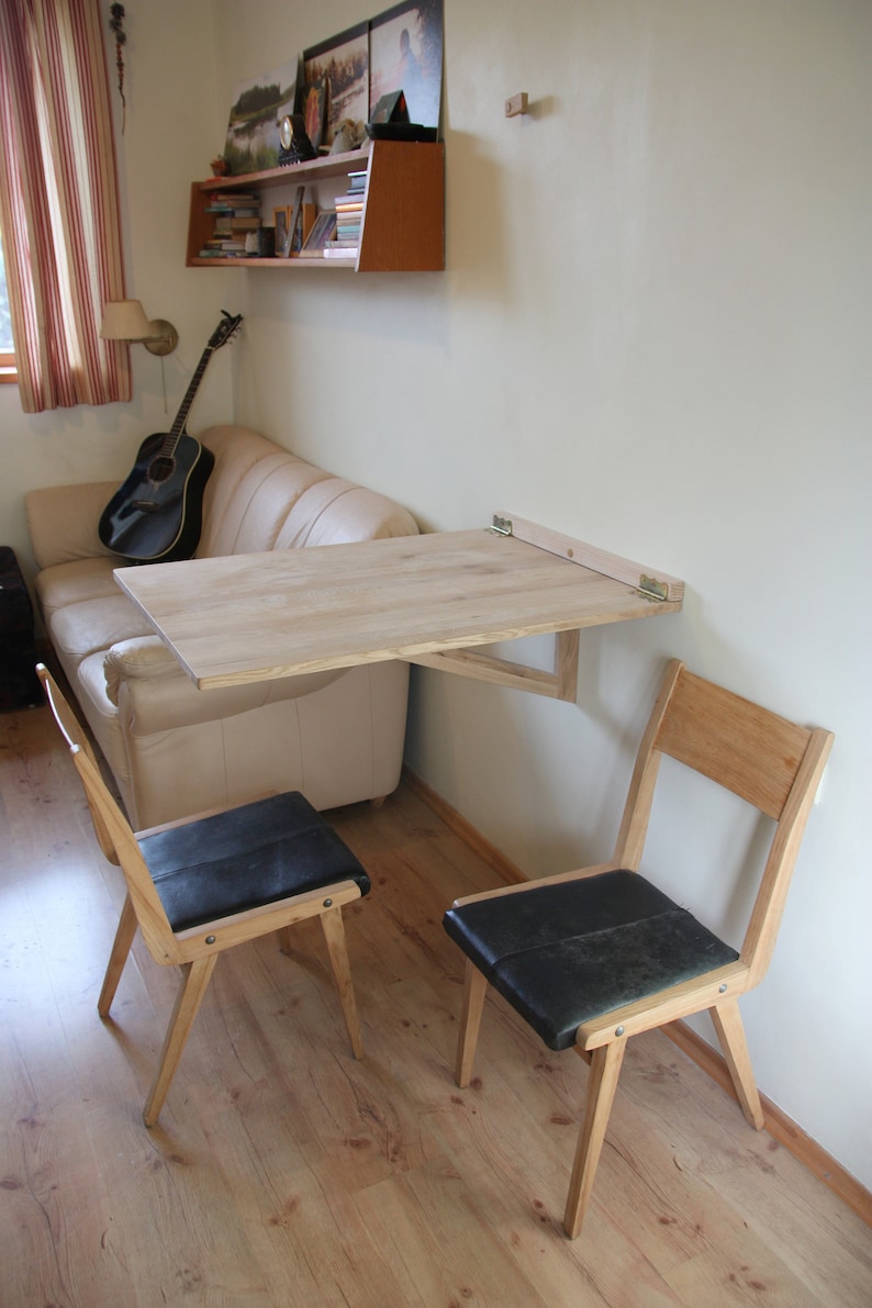 Puede incluir: Una mesa plegable de madera clara con sillas negras est&aacute; montada en una pared. La mesa est&aacute; plegada y no est&aacute; en uso. Las sillas son de madera clara con asientos de cuero negro.