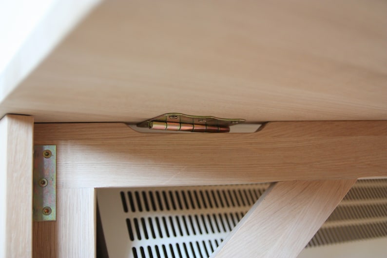 May include: Close-up of a wooden table with a metal hinge connecting the tabletop to the base. The hinge is visible from underneath the table.