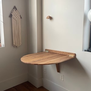 May include: A light wood folding wall table with a rounded edge and brass hinges. The table is mounted to a white wall with a white electrical outlet below.