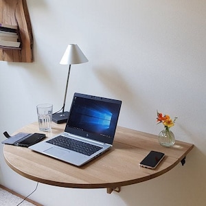 K&ouml;nnte beinhalten: Ein klappbarer Wandtisch aus hellem Holz mit einem Laptop, einem Glas Wasser, einem Handy und einer Lampe. Der Tisch ist an einer wei&szlig;en Wand befestigt.