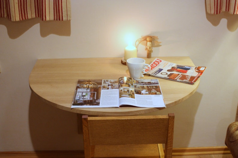K&ouml;nnte beinhalten: Ein heller Holztisch mit abgerundeter Kante, ein aufgeschlagenes Magazin auf der Oberfl&auml;che, eine wei&szlig;e Tasse und eine Kerze auf einem Holzsockel.