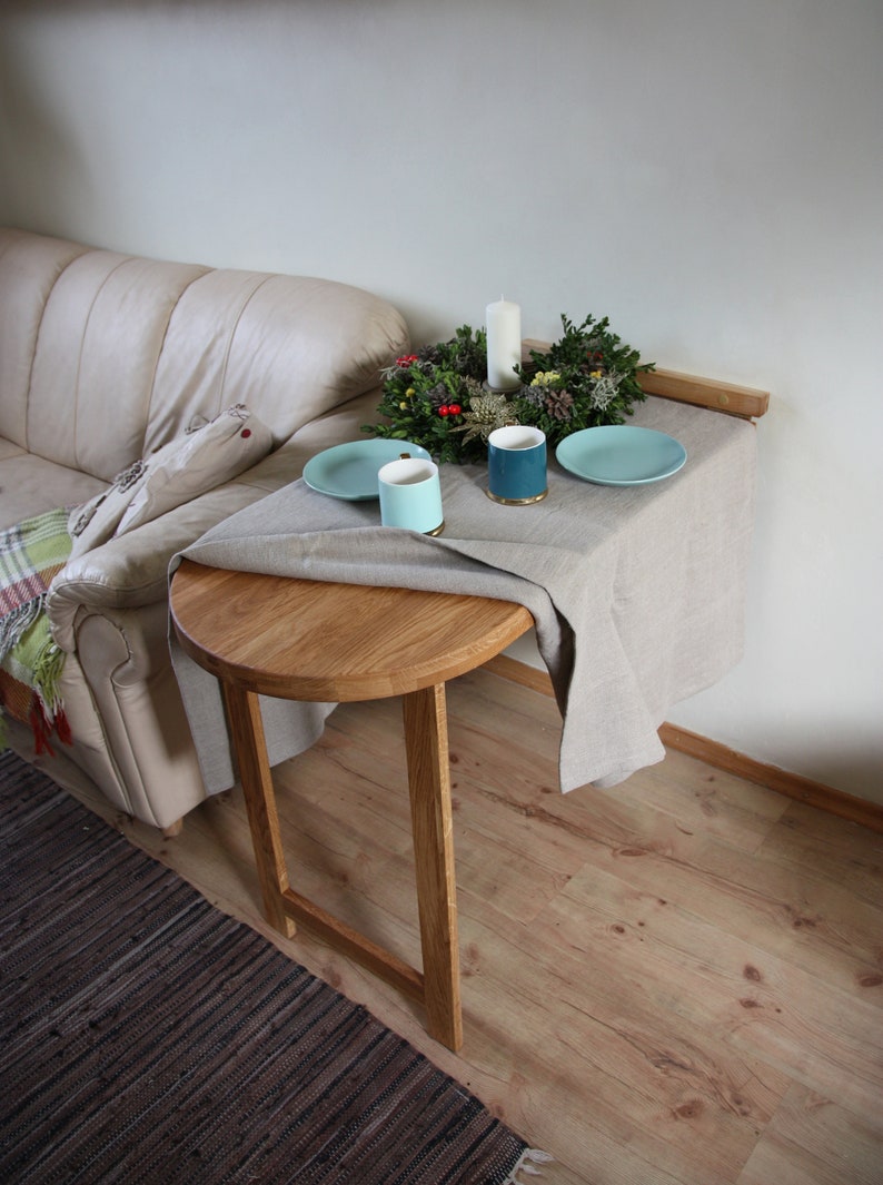 May include: A round wooden table with a light brown finish, set with two blue plates, two blue mugs, and a white candle in a green wreath. The table is attached to a wall and has a linen tablecloth.