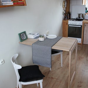 May include: A small, light wood dining table with a drop leaf, paired with a white chair with a black cushion. A black and white patterned table runner and a framed picture are on the table. The table is 30 inches long.