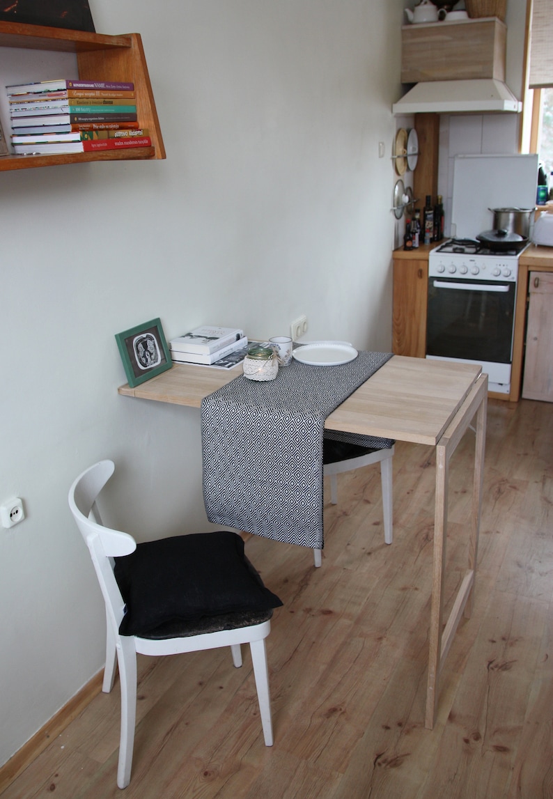 May include: A small, light wood dining table with a drop leaf, paired with a white chair with a black cushion. A black and white patterned table runner and a framed picture are on the table. The table is 30 inches long.