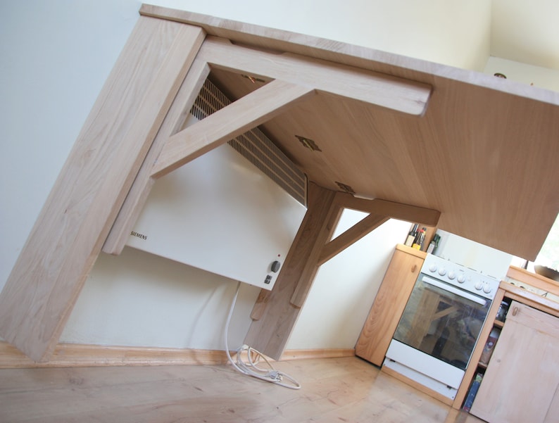 May include: A wooden table with a white electric heater mounted underneath. The table is made of light wood and has a simple design. The heater is a standard model with a white finish.