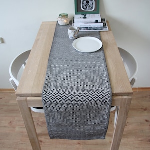 May include: A wooden table with a black and white geometric patterned table runner. A white plate, a floral patterned mug, and a small framed picture are on the table. A small candle and books are also on the table.