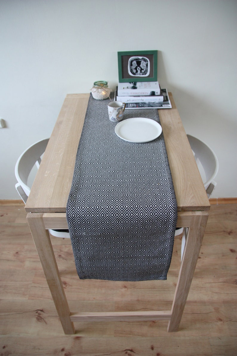 May include: A wooden table with a black and white geometric patterned table runner. A white plate, a floral patterned mug, and a small framed picture are on the table. A small candle and books are also on the table.